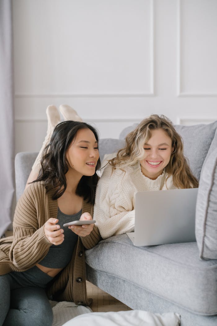 Two women laughing while using a laptop and smartphone in a cozy living room setting.